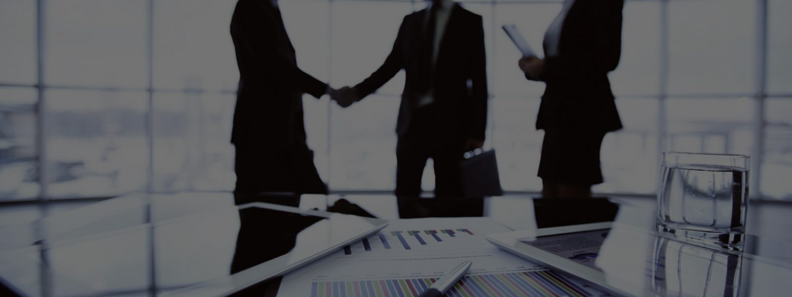 Technological devices, financial document with pen, glass of water at workplace on background of three business partners striking deal